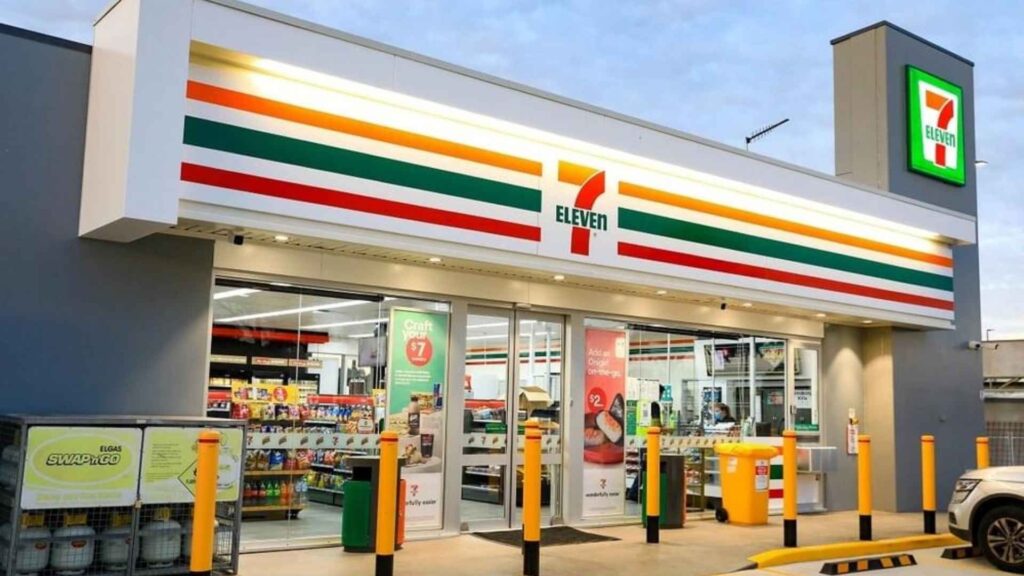 A modern 7-Eleven convenience store building from an outdoor perspective. The storefront is clean and well-lit, with large glass windows and an automatic door. The brand's signature green, red, and orange stripes are visible above the entrance. A yellow "7-Eleven" logo with a red "eleven" is prominently displayed. Signs are visible in the windows, and a yellow recycling bin and bollards are in front of the store. A small part of a vehicle is visible on the far right. The sky is partly cloudy and bright.
