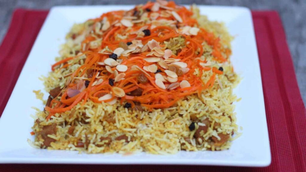 A close-up, top-down shot of a large serving of traditional Afghan Kabuli Pulao on a white platter, garnished with julienned carrots, raisins, and slivered almonds.