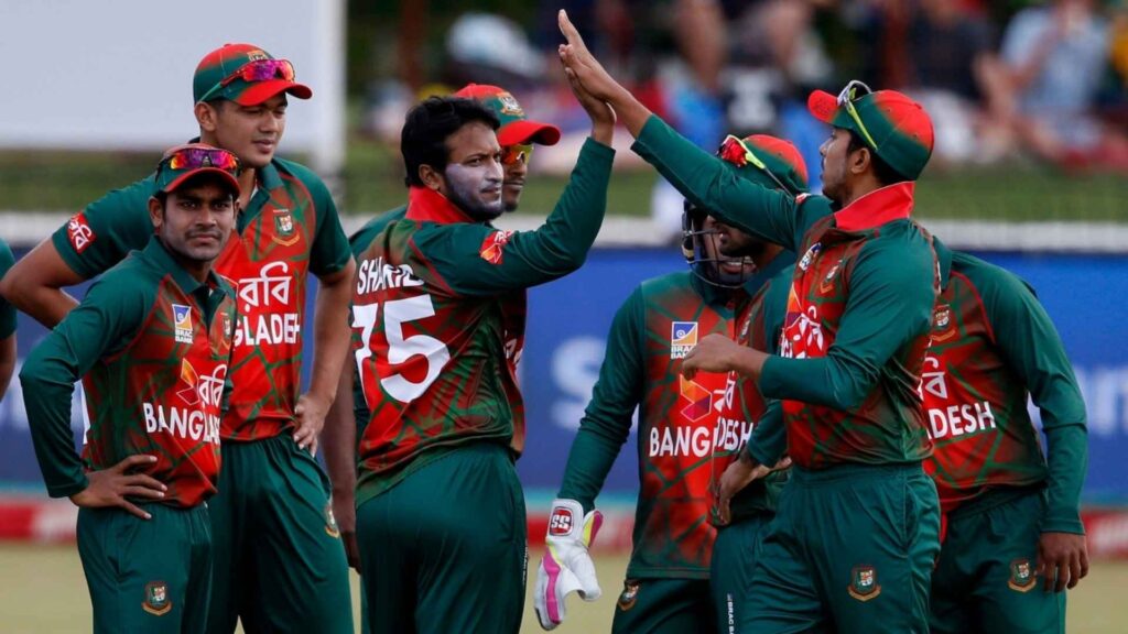  A group of male cricketers from the Bangladesh national team, wearing green and red uniforms, celebrating a play on the field with high-fives and smiles. The player in the center has face paint on his cheeks.