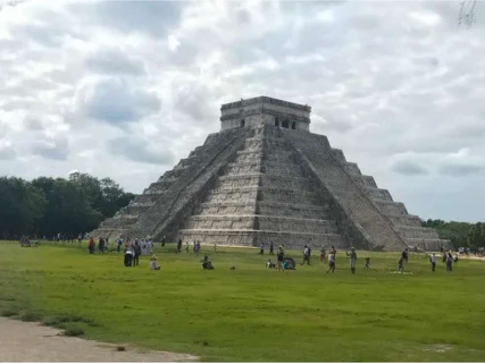 The ancient Mayan step-pyramid, El Castillo (Temple of Kukulcán), at the archaeological site of Chichén Itzá, Mexico. The massive stone pyramid stands on a wide green lawn, with many tourists scattered across the base and trees lining the edges under a cloudy sky.

