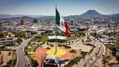 An aerial view of the city of Chihuahua, Mexico, showing a large circular plaza or glorieta with a towering flagpole flying a huge Mexican flag (green, white, and red with the coat of arms). The city's modern and low-rise buildings surround the traffic circle, with dry, rugged mountains visible in the distance under a bright blue sky.

