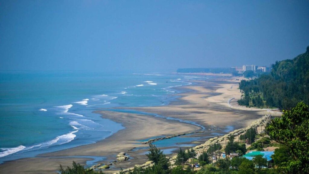  An elevated, wide shot of a vast, sandy beach curving along a calm turquoise sea under a clear blue sky. On the right, a lush green hillside with trees slopes down to the beach, with some buildings visible in the distance.