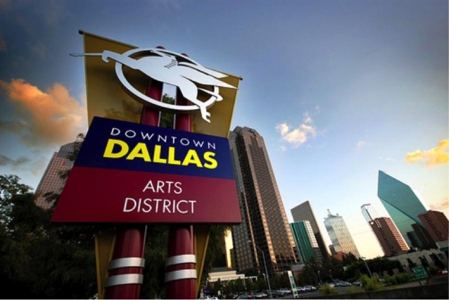 A low-angle shot of a large, colorful sign for the Downtown Dallas Arts District. The sign is made of blue and red panels with yellow and white text. It features the Dallas Pegasus logo on top. In the background, several modern skyscrapers are visible against a cloudy sky with the warm glow of sunset.