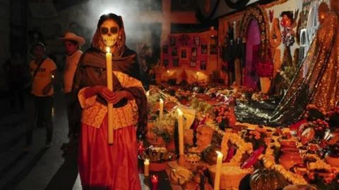 A woman wearing La Catrina skull makeup and traditional clothing, holding a lit candle in a dark, smoke-filled street. Behind her is a large, colorful Day of the Dead (D 
ı
ˊ
 a de Muertos) altar covered in marigold flowers, candles, offerings, and photos of the deceased.

