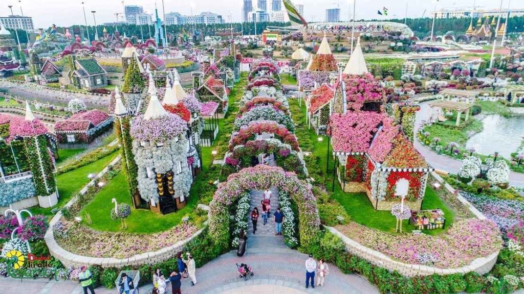 An aerial view of the Dubai Miracle Garden, a large floral park with intricate designs. The image shows several small, house-like structures covered entirely in a dense array of colorful flowers, with paths and archways made of blooms connecting them. In the background, taller buildings and structures are also decorated with flowers.