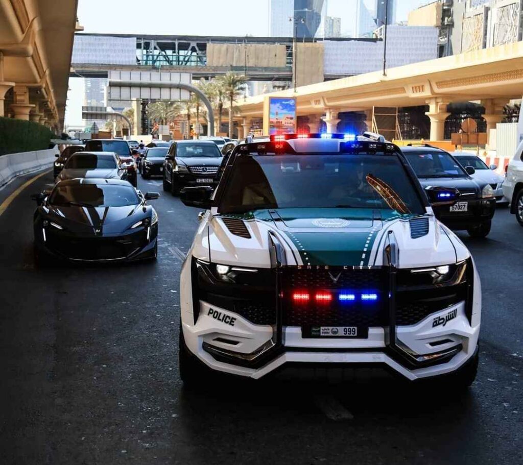 A futuristic-looking, white and green Dubai Police vehicle with flashing red and blue lights drives on a city street, with a black sports car visible beside it and skyscrapers in the background.