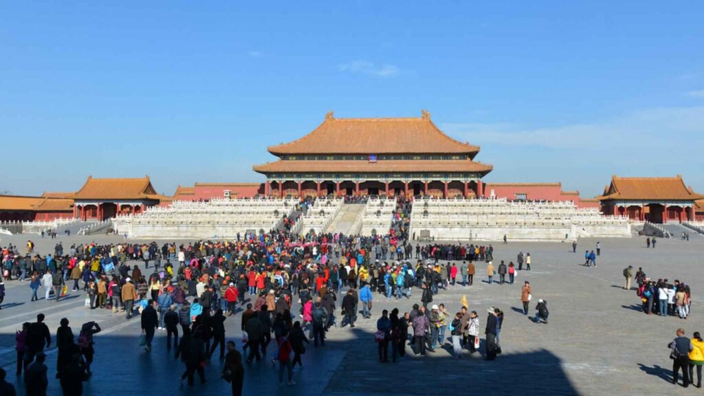 
An expansive view of the Forbidden City's massive courtyard, filled with a large crowd of tourists. In the background, the grand Hall of Supreme Harmony rises on a tiered marble platform, with its distinctive orange-tiled roof and red columns.