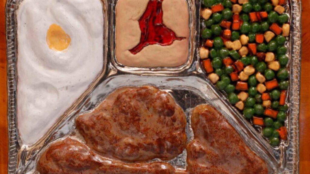 An artistic and realistic representation of a classic frozen TV dinner in a partitioned aluminum tray. The tray contains a main section with two pieces of browned meat, one appearing to be a chicken thigh or drumstick. The three smaller sections contain a white, creamy substance, possibly mashed potatoes with a pat of butter, a square of a pale food with a red gelatinous center, and a mixture of green peas and orange carrots.