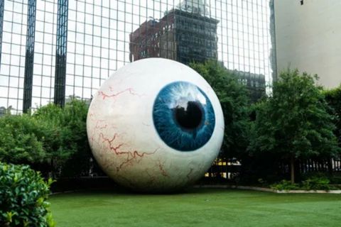 A close-up of the giant eyeball sculpture in Dallas, Texas. The sculpture is a large, hyper-realistic white eyeball with a striking blue iris and red veins. It sits on a patch of green grass, partially surrounded by manicured bushes, with a glass skyscraper and other buildings visible in the reflective background.