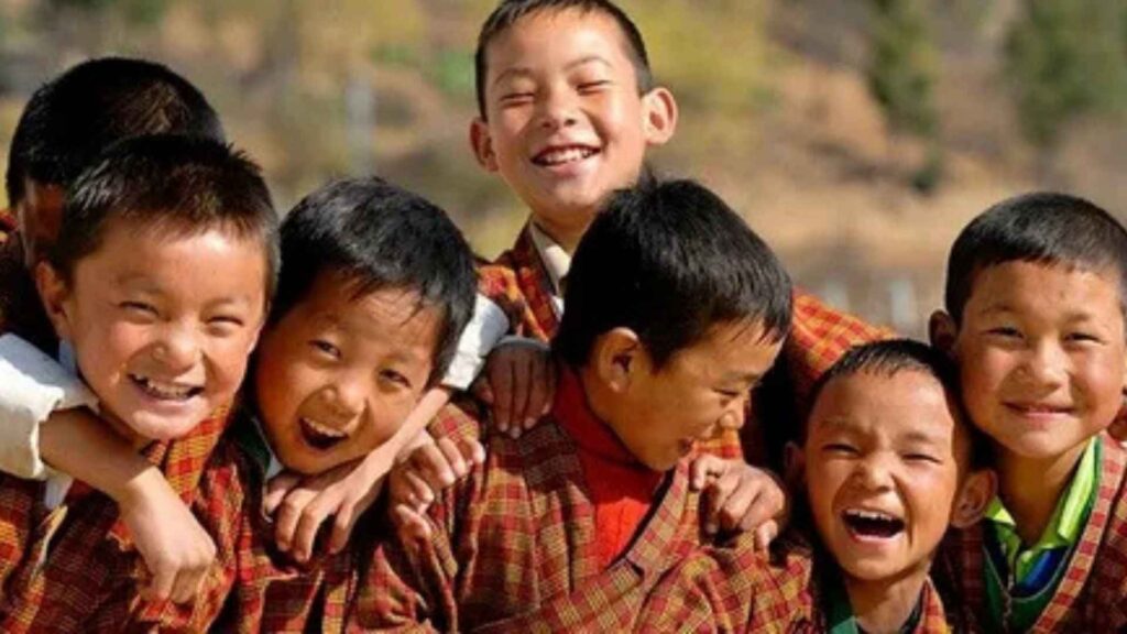 
A group of seven young Bhutanese children, likely school-aged, are gathered together outdoors, smiling and laughing at the camera. They are all wearing traditional Bhutanese clothing, likely a variation of a gho or school uniform, with a plaid or checkered pattern in red, orange, and brown tones. They are standing close together, with some of them embracing each other, conveying a sense of genuine joy and camaraderie. The background is slightly blurred and sun-drenched, suggesting a warm and pleasant day.