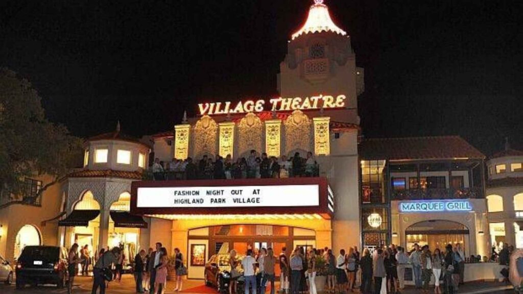 A nighttime photo of the historic Highland Park Village, a luxury shopping center in Dallas, Texas. The prominent "VILLAGE THEATRE" sign is lit up at the top, and a large marquee below reads "FASHION NIGHT OUT AT HIGHLAND PARK VILLAGE." A crowd of people is gathered outside the entrance, with several dressed up for a formal event. The buildings have a distinct Spanish Colonial Revival architectural style, with a tower featuring a red-lit top.