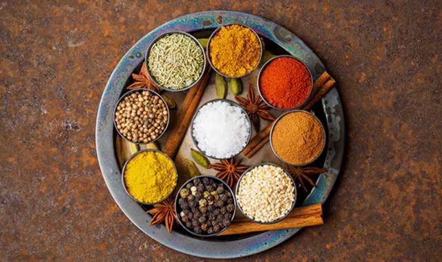 An overhead shot of a round, rustic metal tray holding a variety of small bowls filled with different Indian spices, including turmeric, paprika, black peppercorns, coriander seeds, and sea salt crystals, arranged with cinnamon sticks and star anise.