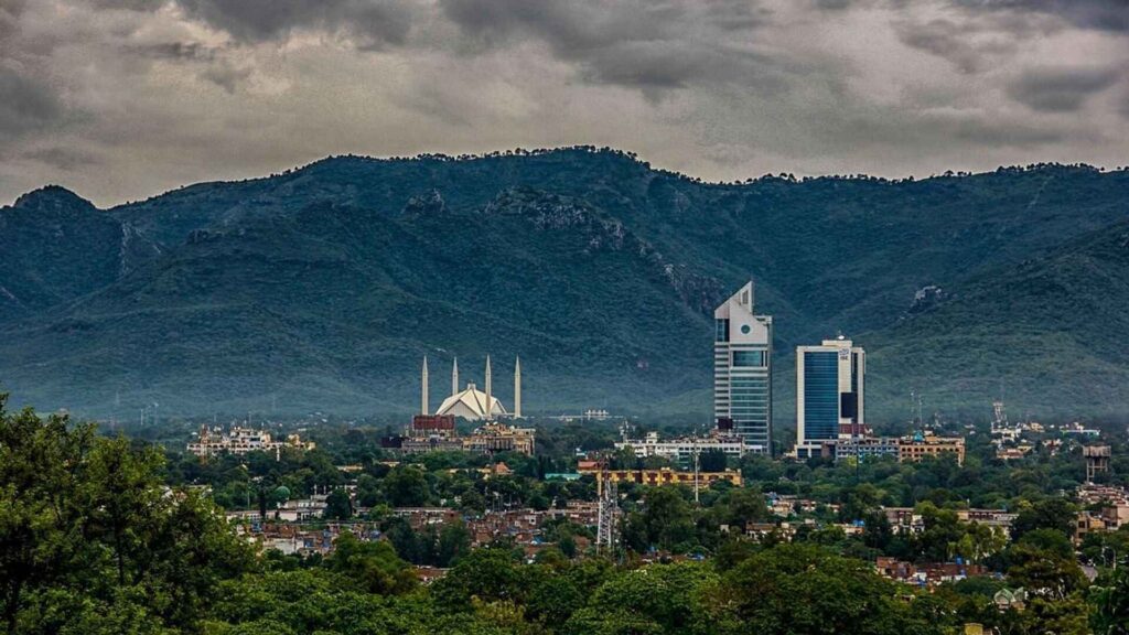 A cityscape of Islamabad, Pakistan, with the Faisal Mosque and modern buildings in the foreground, set against the backdrop of the lush, green Margalla Hills under a dramatic, cloudy sky.