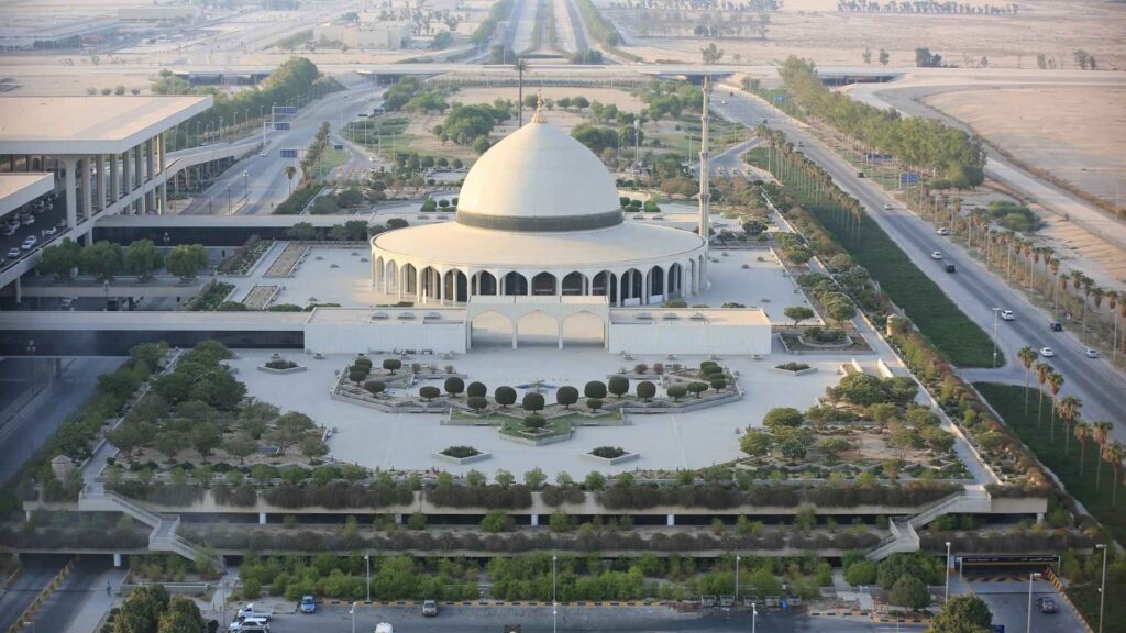 An aerial view of the King Fahd International Airport mosque, a large, modern white mosque with a single large dome and archways, surrounded by meticulously manicured gardens and parking lots. Highways and airport infrastructure are visible in the background, extending into the arid landscape under a hazy sky.

