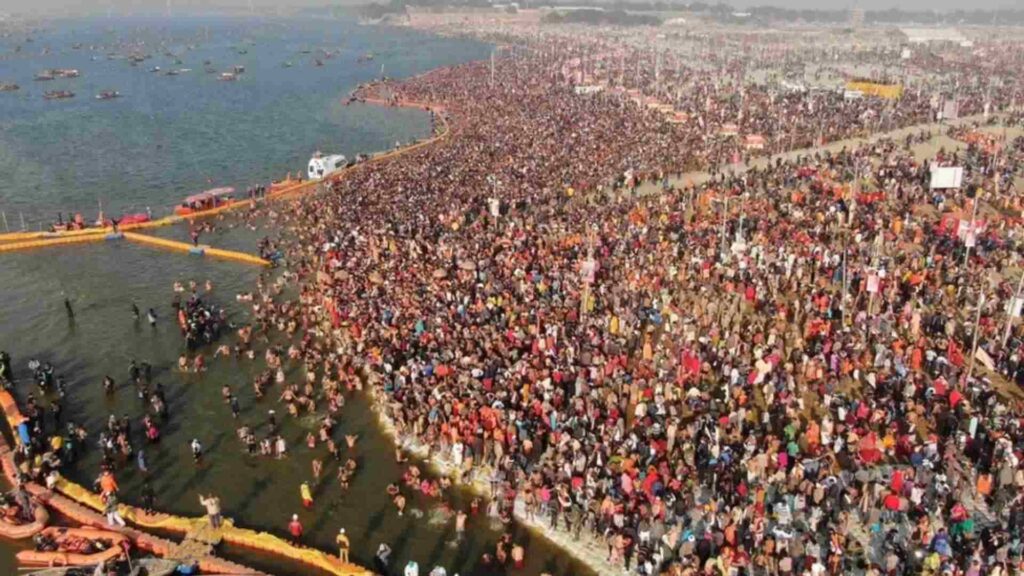 An aerial shot of an extremely dense crowd of people gathered on the banks of a wide river, with many individuals wading and bathing in the water. Numerous small boats are visible in the distance on the river.