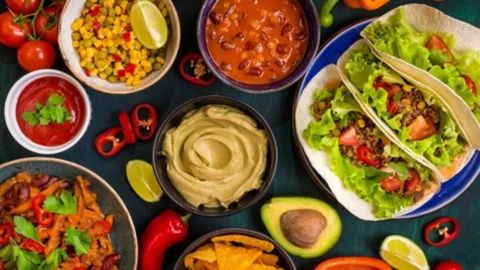 A colorful, overhead flat lay of various Mexican or Tex-Mex dishes, including two ground beef and lettuce tacos in soft tortillas, a bowl of guacamole in the center, chili with beans, corn and pea salsa with lime, a bowl of red salsa, and a plate of nachos, surrounded by whole chili peppers, avocado, and lime slices.










