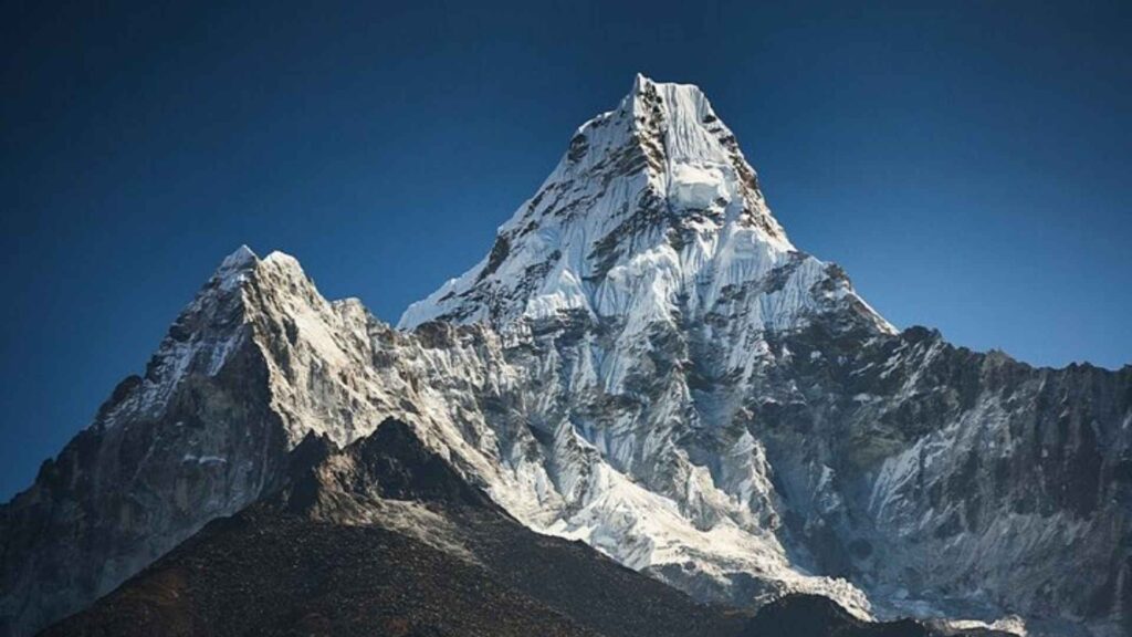 
A sharp, jagged snow-covered mountain peak rises dramatically against a clear blue sky. The lower slopes are rocky and brown.