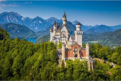 
A horizontal shot of Neuschwanstein Castle, a famous fairytale-like castle in Bavaria, Germany. The castle is perched on a rocky cliff, surrounded by a lush green forest of pine and deciduous trees. The castle's main structure features a light-colored central tower and a facade with red and beige stonework, topped with dark roofs and multiple turrets. In the background, beyond the forested hills, a serene blue lake is visible, with the towering, jagged peaks of the snow-capped Alps filling the horizon under a clear blue sky.