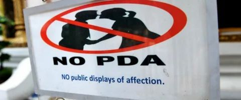 
A rectangular white sign with a red circle and slash symbol over a silhouette of two people kissing. The sign reads "NO PDA" in large black letters and "NO public displays of affection." in smaller blue letters below.