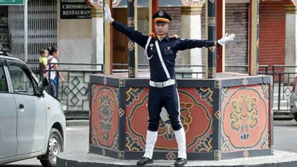 A Bhutanese traffic police officer, dressed in a traditional blue uniform with white gloves, stands on a decorated podium in the middle of a street intersection. He has his arms outstretched, directing traffic. A car is visible on the left side, and a traditional building with a restaurant sign is in the background. The officer's pose is formal and precise, highlighting the unique way traffic is managed in Thimphu without traffic lights.