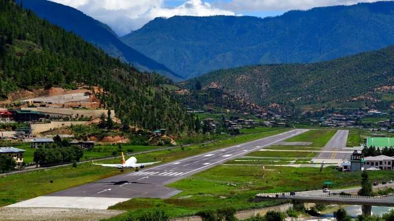 A wide-angle aerial or elevated shot of Paro Airport in Bhutan. An airplane, possibly a Drukair aircraft, is captured just moments before landing on a single runway that stretches through a narrow valley. The airport is surrounded by steep, green, forested mountains on all sides, with a river flowing near the landing strip. Traditional Bhutanese buildings with distinctive architecture are visible on the airport grounds, and the overall scene highlights the challenging yet picturesque nature of the landing approach.









