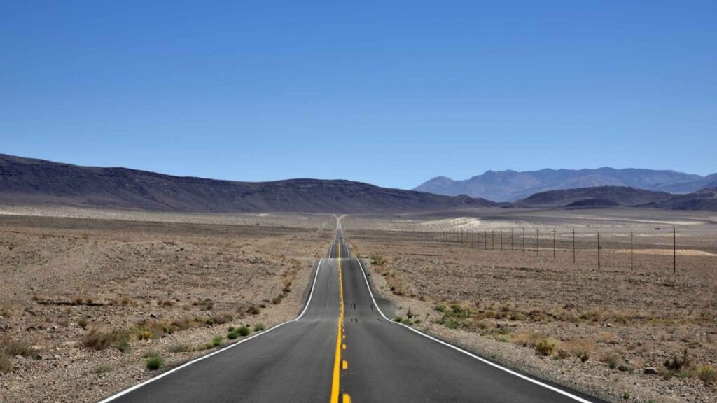: An extremely long, straight road with double yellow lines in the middle extends toward the horizon through a vast, arid desert landscape. Low, brown mountains are visible on both sides under a clear, bright blue sky.