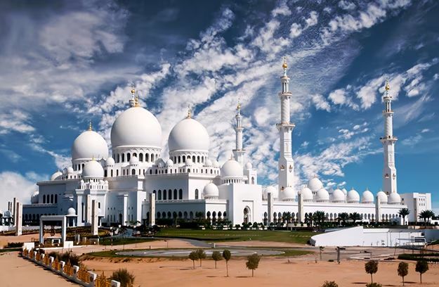A low-angle shot of the Sheikh Zayed Grand Mosque in Abu Dhabi, a massive white marble structure with a large central dome and several smaller domes and minarets, set against a dramatic sky with scattered clouds