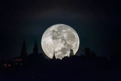  A large, luminous full moon hangs in a dark night sky behind the silhouette of a Buddhist stupa and trees on a hill. The stupa's spire is visible just below the moon, creating a dramatic and serene scene.