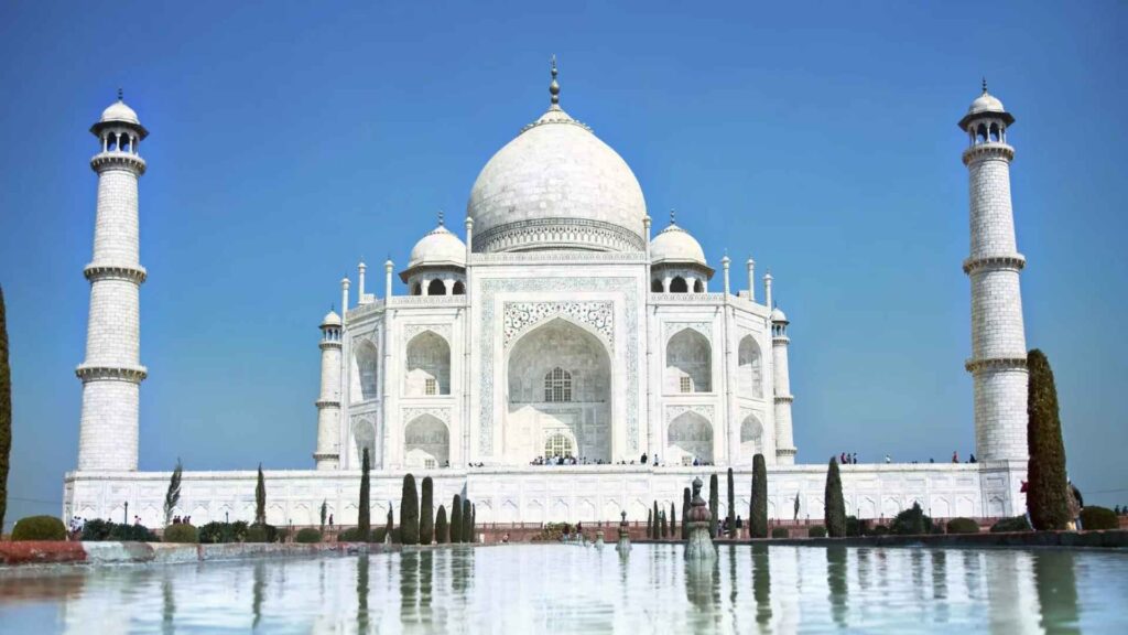 A straight-on, low-angle shot of the Taj Mahal, a magnificent white marble mausoleum with a large central dome and four surrounding minarets, reflected in a long pool of water. The sky above is a clear, brilliant blue.