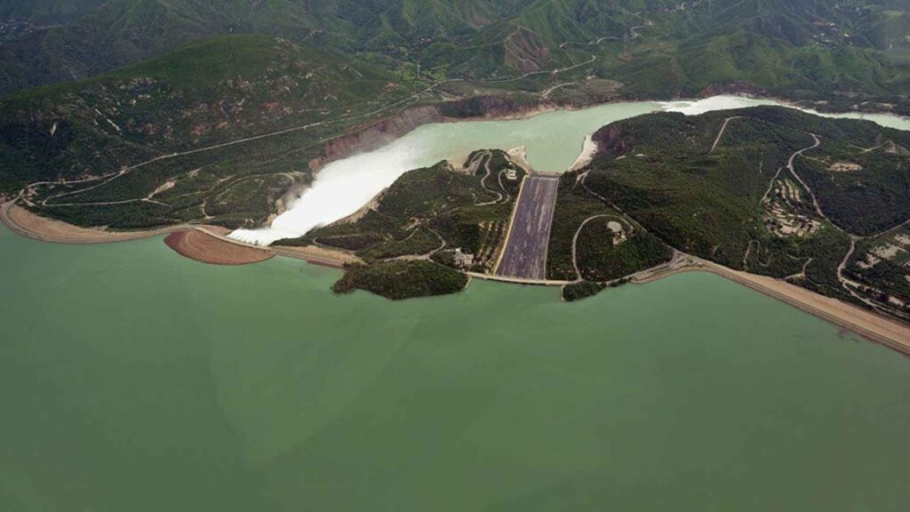 Aerial view of Tarbela Dam in Pakistan with water flowing through the spillways and surrounded by lush green hills.