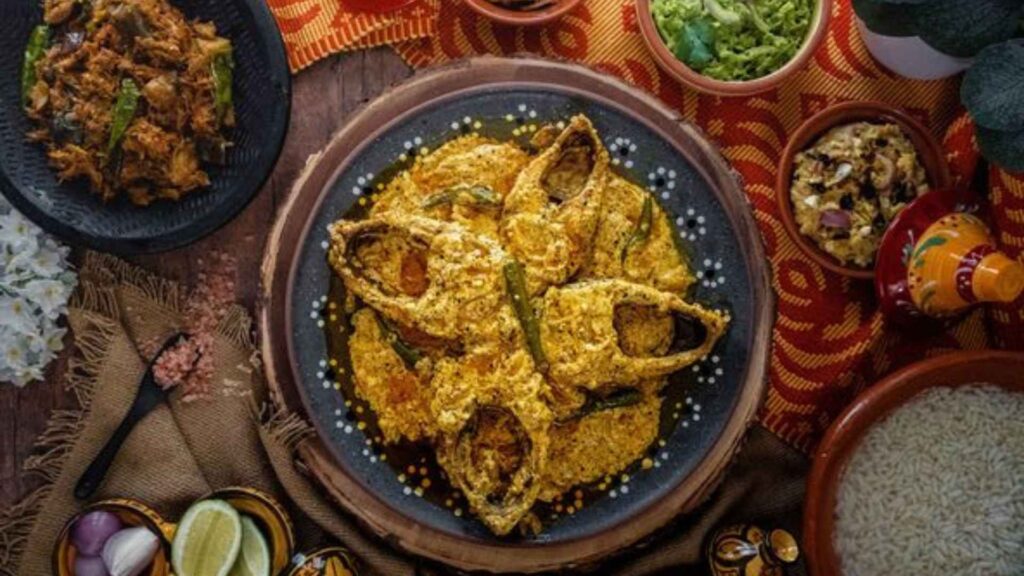 An overhead shot of a Bangladeshi feast featuring a large platter of fried hilsa fish in a rich, yellow curry sauce, surrounded by various bhorta and  side dishes.