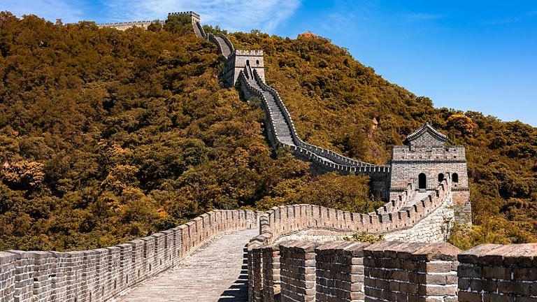 A scenic view of a section of the Great Wall of China, a stone path winding up and down over a forested hillside with several watchtowers.