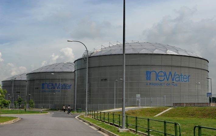 
The image shows three large, cylindrical water tanks with domed roofs, set against a partly cloudy sky. The tanks are gray and have the words "neWater" and "A PRODUCT OF PUB" printed on their sides in blue. They appear to be part of a large-scale water treatment facility, with a paved road and a grassy area with a fence in the foreground.