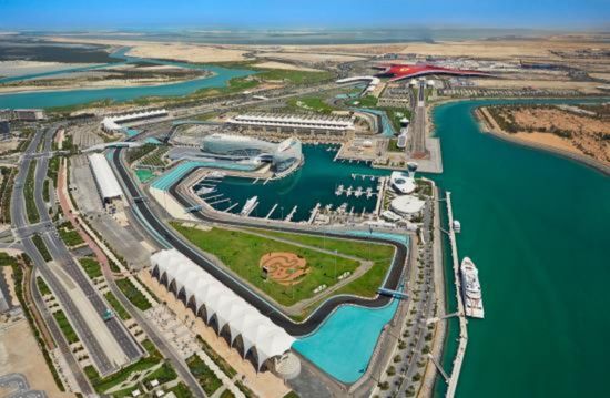 An aerial view of the Yas Island complex in Abu Dhabi, showing the Yas Marina Circuit, a modern marina with yachts, and the red Yas Island theme park building in the distance. The area is surrounded by water and undeveloped land.