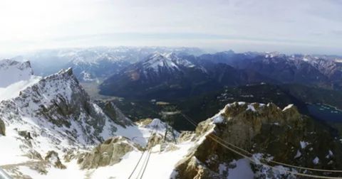 A breathtaking panoramic view from the summit of a snow-covered mountain, likely the Zugspitze in the German Alps. In the foreground, the jagged, rocky peaks and snow-dusted slopes of the surrounding mountains are visible. A few cable car lines stretch across the frame, leading down into the valley below. The expansive middle and background show a vast landscape of majestic mountain ranges, some with patches of snow, stretching out to the horizon under a bright, hazy sky. A small, dark blue lake can be seen in the distance on the right.
