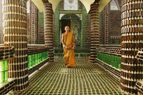 
An orange-robed Buddhist monk stands centered in the covered walkway of the Wat Pa Maha Chedi Kaew (Temple of a Million Bottles) in Sisaket Province, Thailand. The entire structure, including the floor, pillars, and railings, is constructed from thousands of green and brown glass bottles laid on their sides to create a striking mosaic pattern. The monk is facing the camera with his hands clasped in front of him, creating a strong vertical element that contrasts with the textured glass walls.