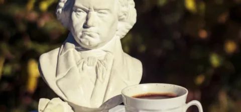 A white bust statue of the composer Ludwig van Beethoven positioned outdoors, with a white mug of black coffee sitting on a saucer in the foreground. beautiful coffee facts