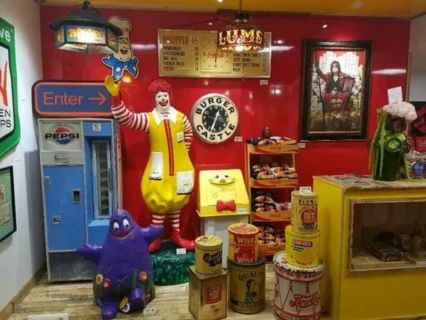 A display of vintage fast-food and soda memorabilia, including a life-sized statue of Ronald McDonald, a purple Grimace statue, a Pepsi vending machine, and various retro signs, clocks, and canned goods. The items are arranged against a bright red wall, giving the appearance of a fast-food museum or collection.