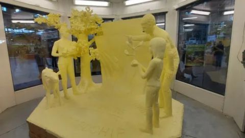 A large, pale yellow butter sculpture displayed under glass in an exhibit hall. The sculpture features a scene depicting human and animal figures—specifically an adult male and two younger figures (a boy and girl)—alongside a tree and a small cow. The entire piece rests on a platform that has the word "LEADERSHIP" carved into the front.