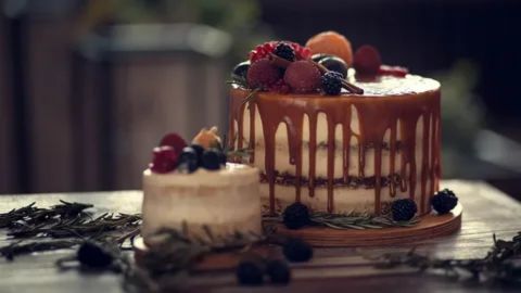 A close-up photograph of four small, decadent layered cakes arranged on a round wooden serving board. The cakes vary in flavor and decoration: one is white with a chocolate drizzle and strawberries, another is chocolate with a heavier chocolate ganache drizzle and strawberries, a third is a chocolate layer cake with a thick white filling and whipped cream, and the fourth is a solid dark chocolate cake topped with a caramel drizzle and gold accents. All cakes are garnished with fresh, whole strawberries or chocolate shavings.