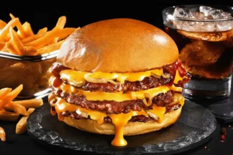 A large, stacked double cheeseburger with melted cheddar cheese dripping down the side of the bottom bun, topped with caramelized onions. The burger is presented on a dark, slate surface next to a wire basket overflowing with golden french fries and a glass of dark soda with ice.
