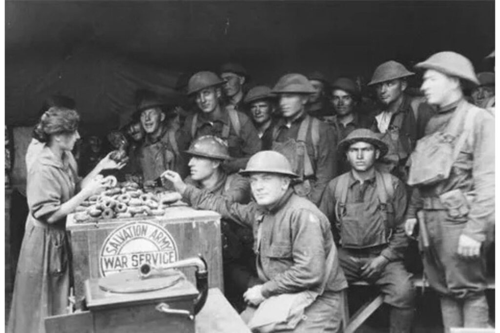 A black-and-white photo from World War I. A woman in a dress and apron, identified as part of the Salvation Army War Service by a sign on the counter, is handing donuts to a line of smiling American soldiers in uniform and helmets. A large pile of donuts is visible on the wooden counter.