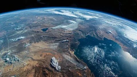An orbital view of the Earth's surface, dominated by a mix of brown, mountainous terrain and desert areas, with patches of snow or white clouds over a large landmass. A large blue sea or gulf is visible on the right, contrasting with the detailed, rugged land. The curve of the planet's horizon and the blackness of space are visible at the top.