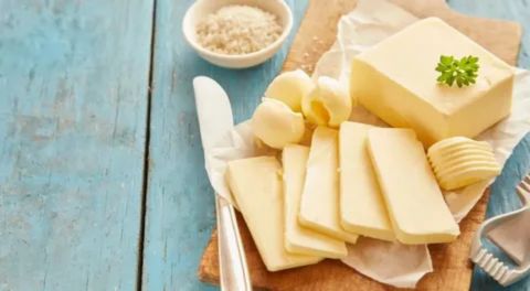 A high-angle shot shows a large block of butter topped with a parsley garnish, several thin butter slices, and small curled butter pats on a wooden cutting board. A white bowl of salt flakes is visible in the upper corner, all resting on a rustic blue wooden surface.