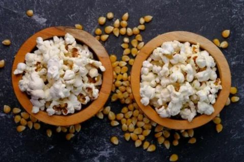 Top-down shot of a dark background featuring two wooden bowls brimming with fresh popcorn, contrasting with the scattered golden-yellow popcorn kernels around them.