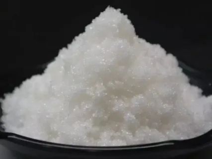 A close-up of a white, crystalline, granular pile of material, likely sodium acetate (commonly known as "hot ice"), resting on a black surface.










