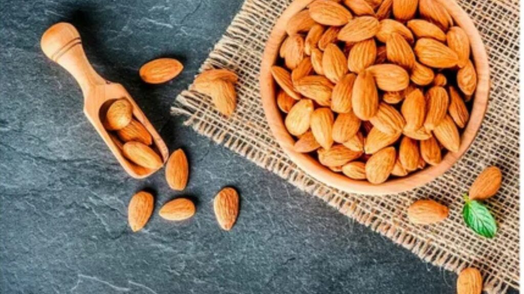 A bowl filled with raw almonds on a grey marble floor with some scattered almonds and wooden spoons beside it having some almonds in it.