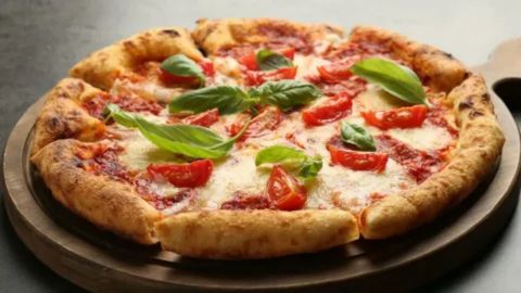 A delicious, close-up, and slightly low-angle photo of a freshly baked Margherita pizza on a rustic round wooden peel or cutting board. The pizza has a thick, charred, artisanal crust and is topped with bright red tomato sauce, melted white mozzarella cheese, large slices of fresh red tomato, and vibrant green basil leaves. The background is dark and blurred, drawing full attention to the colorful ingredients of the classic Italian dish.