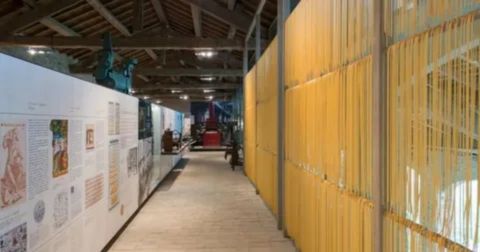 Museum interior showing a long hallway with a wall of vertically hanging, bright yellow dried spaghetti strands on the right and an informational exhibit wall with historical documents and text on the left.

