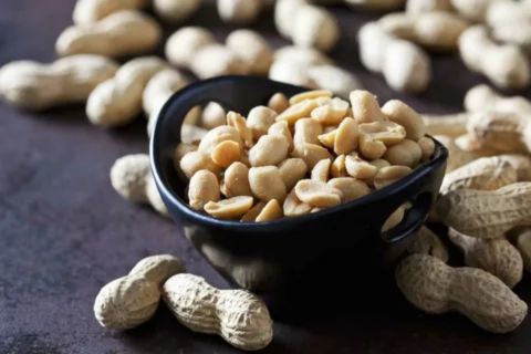 A close-up of a small black bowl filled with shelled, roasted peanuts, surrounded by scattered whole, unshelled peanuts on a dark, textured surface